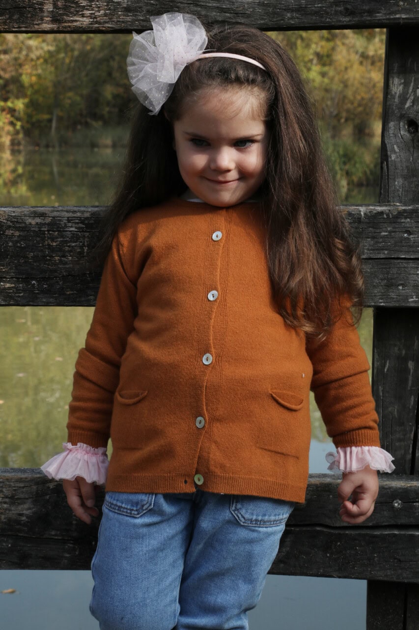 Dolce bambina con cardigan arancione e dettagli raffinati, perfetto per l'autunno, con ambiente naturale sullo sfondo.