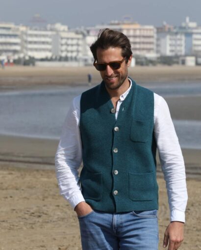 Uomo sorridente indossa un gilet in lana blu con bottoni, camicia bianca e jeans, passeggiando sulla spiaggia con sfondo di edifici balneari e mare.