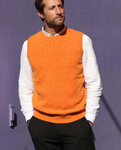 Uomo elegante indossa un gilet arancione in maglia, abbinato a camicia bianca e pantaloni neri, in un ambiente urbano con parete viola sullo sfondo.
