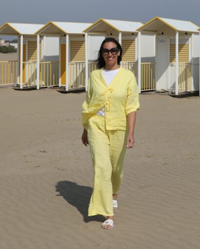 Vestito estivo giallo, donna sorridente in spiaggia con cabine colorate sullo sfondo, perfetto per look casual e alla moda durante le vacanze al mare.
