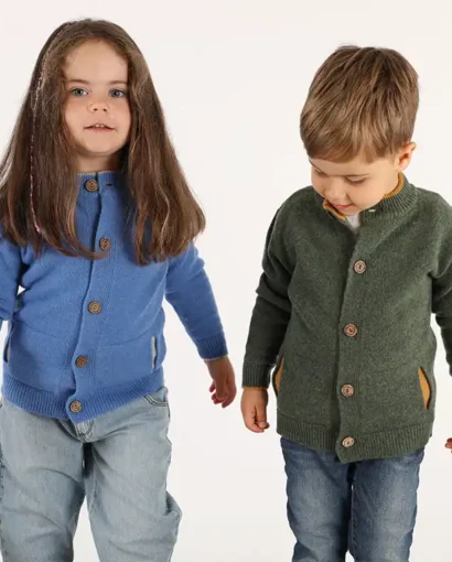 Due bambini indossano cardigan colorati, uno blu e uno verde, in un ambiente neutro, mostrando abbigliamento di alta qualità del Maglificio Annamaria.