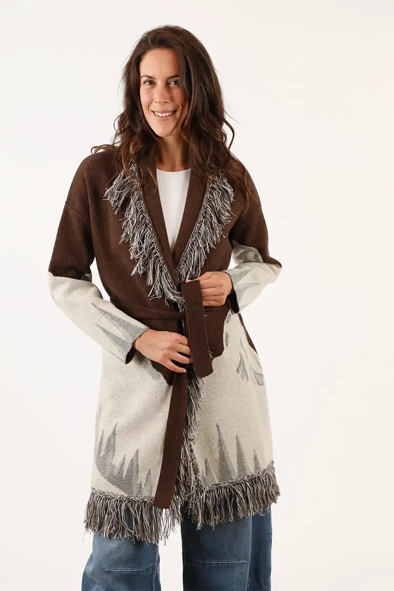 Donna indossa un cappotto lungo in maglia con dettagli frange e colori marrone, beige e grigio, perfetto per l'autunno e l'inverno, rappresentando l'eleganza del Maglificio Annamaria.