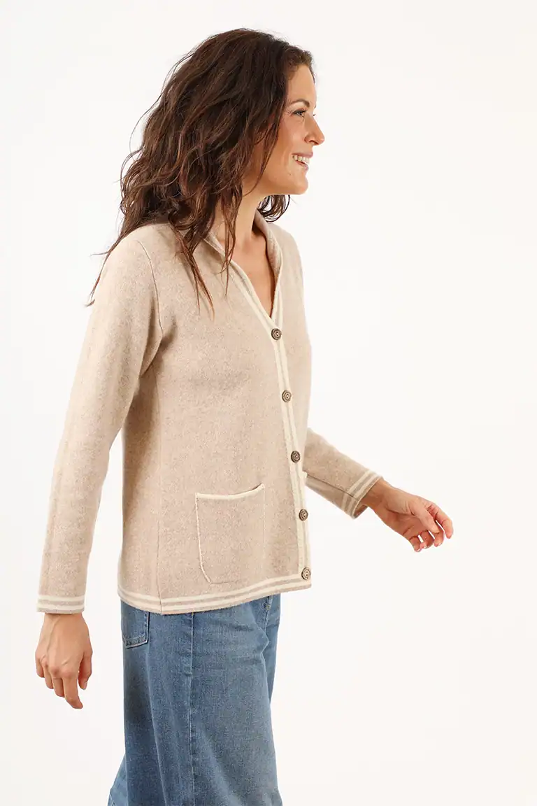 Donna sorridente indossa un cardigan beige con bottoni e tasche, abbinato a jeans blu, in un ambiente neutro, promuovendo moda e maglieria di alta qualità di Maglificio Annamaria.