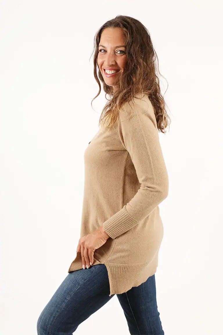 Donna sorridente indossa un maglione beige lungo e jeans, rappresentando l'abbigliamento di alta qualità di Maglificio Annamaria, specializzato in maglieria artigianale italiana.