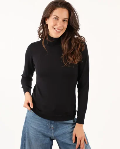 Donna indossa un maglione nero e jeans blu, sorridente, in un ambiente neutro, rappresentando l'eleganza e il comfort dei capi del Maglificio Annamaria.