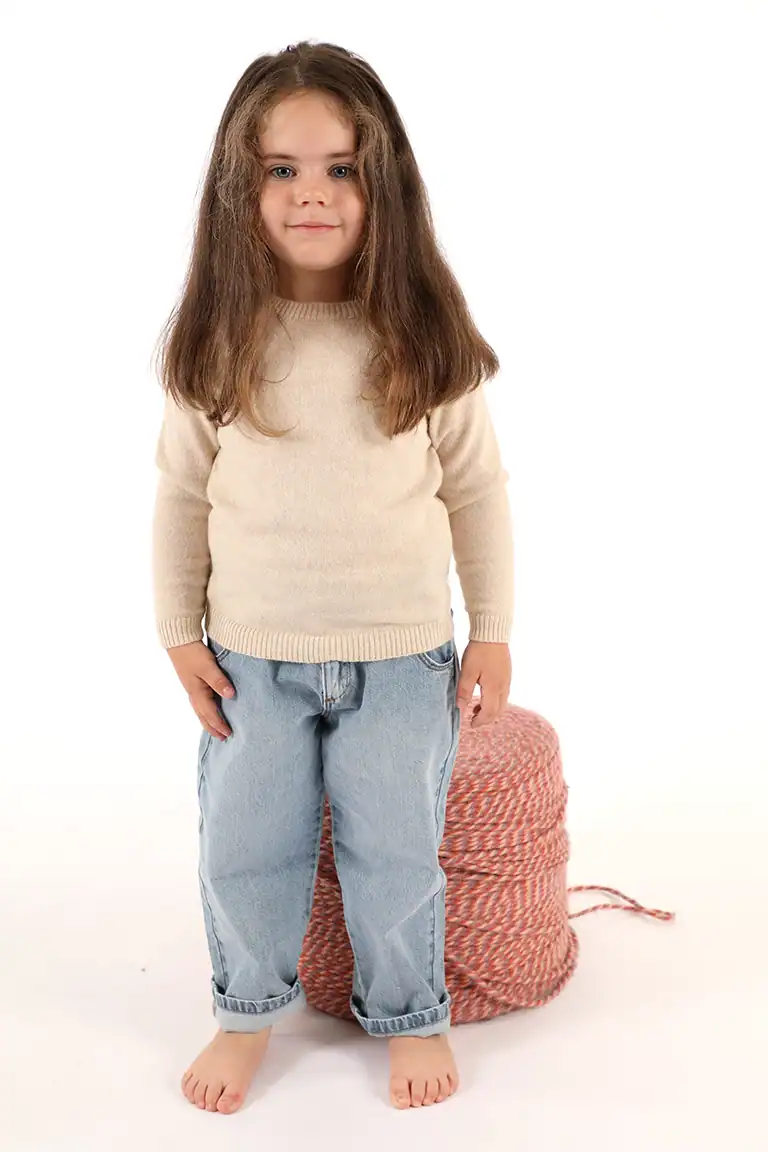 Bambina con maglione beige e pantaloni larghi in denim, posa su sfondo bianco, rappresenta abbigliamento per bambini di alta qualità da Maglificio Annamaria.