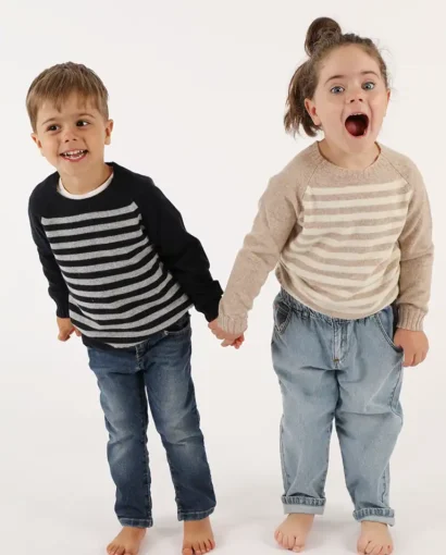 Due bambini felici indossano maglioni a righe e jeans, tenendosi per mano, in uno studio bianco, promuovendo abbigliamento per bambini di Maglificio Annamaria.
