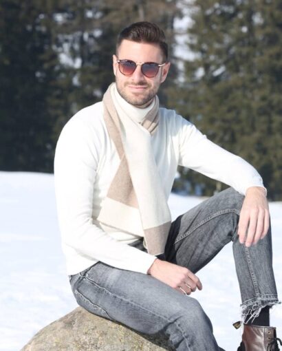 Uomo con occhiali da sole, maglione bianco, sciarpa beige, jeans grigi e stivali in piedi su una roccia in un paesaggio invernale con alberi e neve.