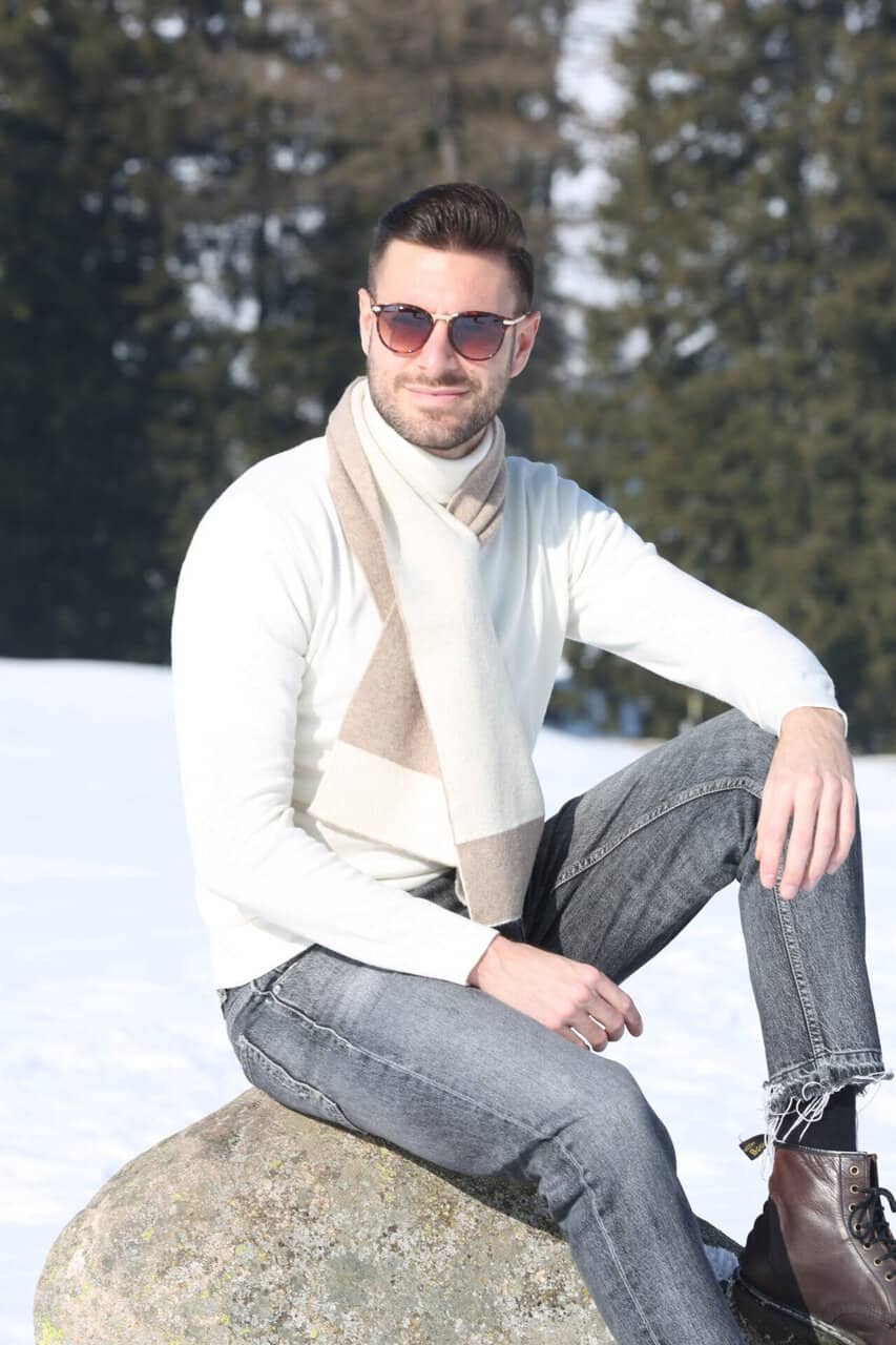 Uomo con occhiali da sole, maglione bianco, sciarpa beige, jeans grigi e stivali in piedi su una roccia in un paesaggio invernale con alberi e neve.