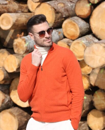Uomo con giacca in maglia arancione, occhiali da sole, sfondo di tronchi di legno, stile casual elegante, moda uomo, maglificio Annamaria.