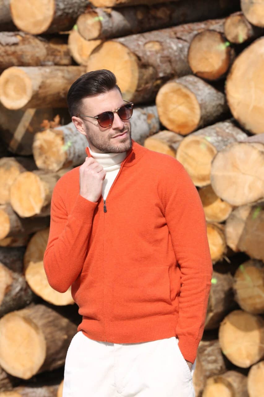 Uomo con giacca in maglia arancione, occhiali da sole, sfondo di tronchi di legno, stile casual elegante, moda uomo, maglificio Annamaria.