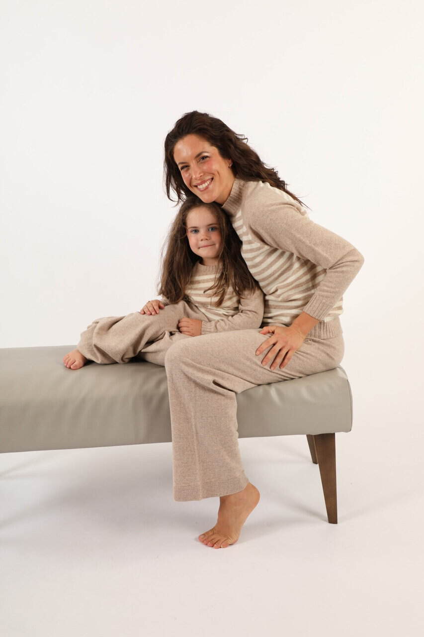Donna e bambina sorridenti indossano maglioni e pantaloni in maglia beige, sedute su una panca bianca in uno studio fotografico per promuovere i capi di Maglificio Annamaria.