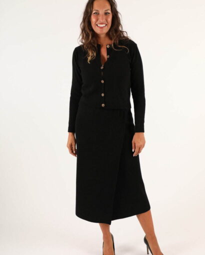 Donna sorridente indossa un completo nero elegante con gonna lunga e cardigan con bottoni, in uno studio bianco, rappresentando moda e stile italiano.