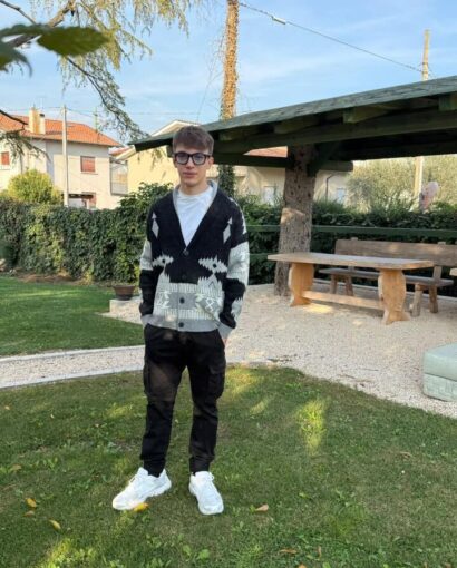 Uomo con maglione a fantasia nero e bianco, pantaloni neri e scarpe bianche in un parco soleggiato, con struttura in legno e case sullo sfondo.